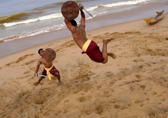 Kalaripayattu acrobatic training on Kerala beach with practitioners performing aerial moves and shield techniques in natural setting